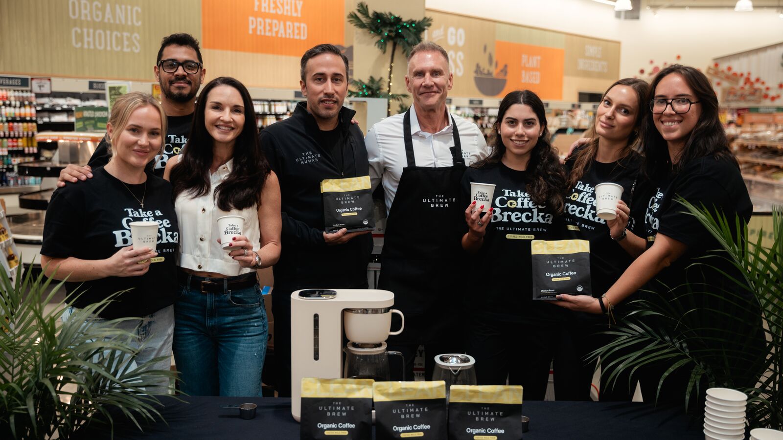 Gary Brecka and the Coffee Brecka team behind the table at Sprouts Coconut Grove, holding bags and cups of The Ultimate Brew