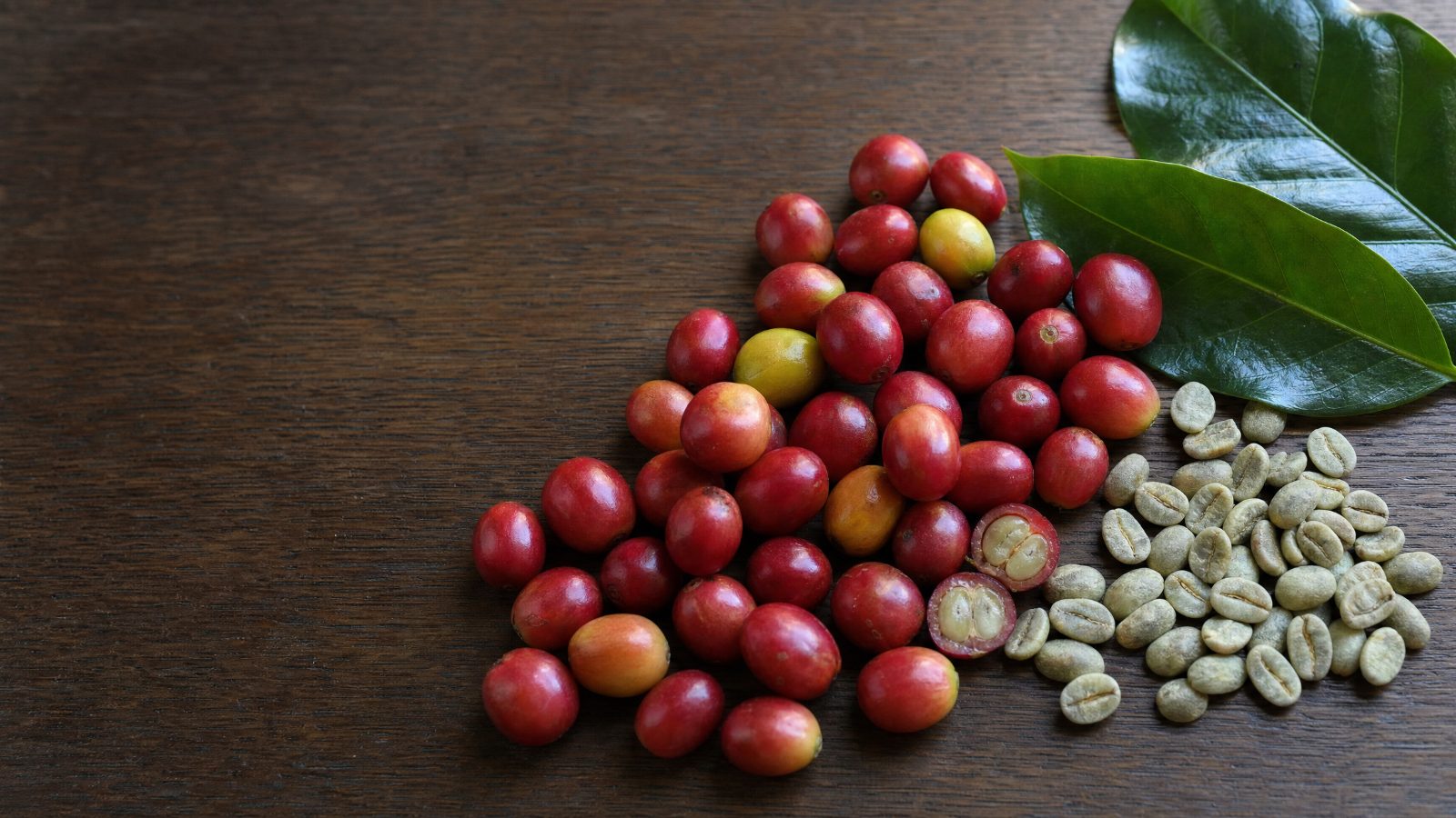 Ripe red and yellow coffee cherries on a dark wood table, one split open to show its seeds, beside a heap of green coffee beans and two coffee leaves